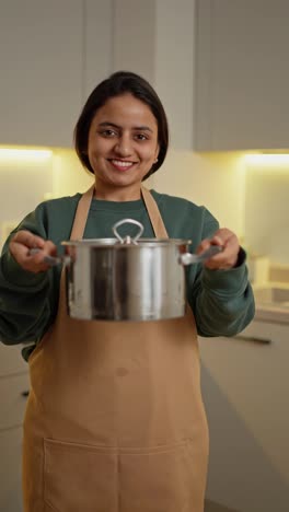 Portrait-of-a-happy-brunette-Indian-girl-in-a-green-sweater-and-beige-apron-holding-a-gray-shiny-pan-in-her-hands-in-a-modern-kitchen-in-a-modern-apartment